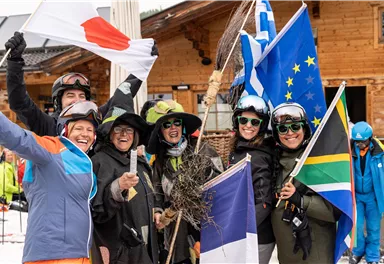 Eine Gruppe von fröhlichen Skifahrern steht zusammen und hält verschiedene Flaggen. Sie tragen Wintersportbekleidung und lächeln in die Kamera.