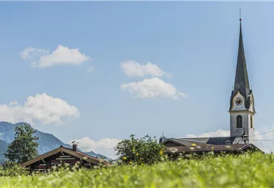 Eine malerische Landschaft mit einer Kirche und einem traditionellem Gebäude im Vordergrund. Der Himmel ist blau mit einigen Wolken und die Berge sind im Hintergrund sichtbar.