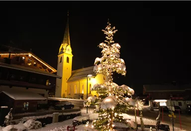 Ein festlich geschmückter Weihnachtsbaum steht im Schnee, beleuchtet von warmem Licht. Im Hintergrund ist eine Kirche mit einem hohen Turm zu sehen, die ebenfalls in warmes Licht getaucht ist.