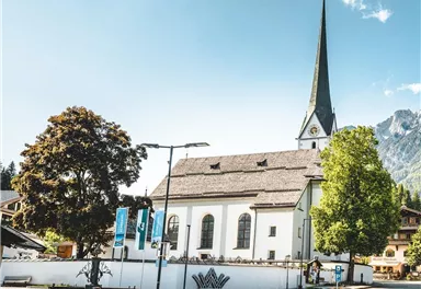 A picturesque church with a tall tower surrounded by trees. In the background, mountains and a clear blue sky can be seen.