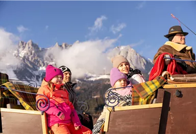 Eine fröhliche Familie auf einer Pferdeschlittenfahrt in den Bergen. Im Hintergrund sind schneebedeckte Gipfel und ein klarer Himmel zu sehen.