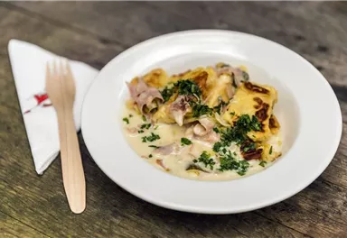 A plate of baked pasta in a creamy sauce, garnished with fresh herbs. Next to it lies a wooden cutlery and a cloth napkin.