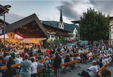 A lively outdoor concert event with many spectators. In the background, there is a church with a tall tower and picturesque mountains.