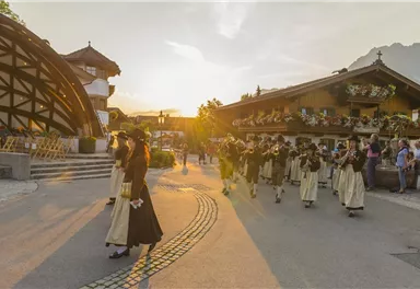 Eine Gruppe von Menschen in traditioneller Kleidung marschiert durch eine malerische Straße. Im Hintergrund sind schöne Holzhäuser und eine sonnige Atmosphäre sichtbar.