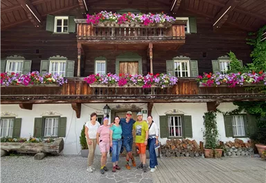 Eine Gruppe von sechs Personen steht vor einem traditionellen, blumengeschmückten Bauernhaus. Die Fassade ist aus Holz und gibt einen einladenden Eindruck.