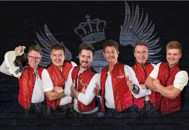 A group of six musicians stands smiling in front of a black wall with a crown and wing motif. They are wearing red vests and display a friendly, inviting demeanor.