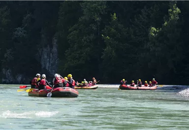 Eine Gruppe von Menschen fährt mit Schlauchbooten auf einem Fluss. Die Umgebung ist grün und bewaldet.