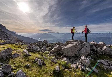 Zwei Wanderer stehen auf einem Felsen in den Bergen und genießen die Aussicht. Der Himmel ist klar mit sanften Wolken und die Landschaft ist grün und bergig.