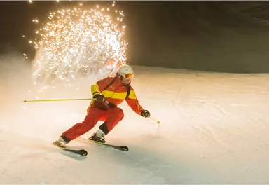 A skier in a red-yellow suit is skiing through the snow. Sparks fly behind him, illuminating the wintry landscape.