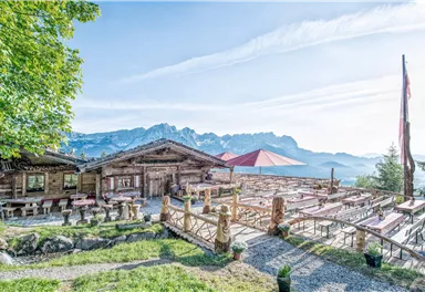 A rustic mountain restaurant with wooden construction and a beautiful outdoor terrace. In the background, majestic mountains and a clear sky can be seen.