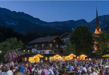 A lively night festival with many people and festively illuminated stalls. In the background, mountains rise and a church with a lit tower.