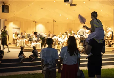 A family is watching a concert on a stage. In the background, a band is playing while the audience enjoys the evening in a relaxed manner.