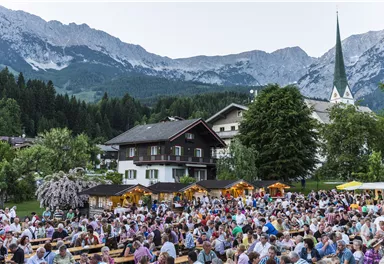 A large crowd sits in a picturesque village among the mountains. Surrounded by trees and traditional buildings, the visitors enjoy a festive atmosphere.