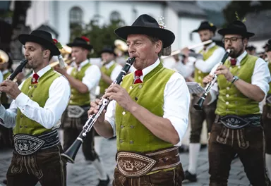 A group of musicians in traditional costumes plays instruments. They are standing in a square and have a cheerful expression.