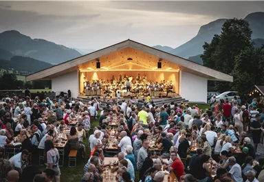A large crowd is enjoying an outdoor concert. A music band is playing on stage, while in the foreground there are many tables and chairs.