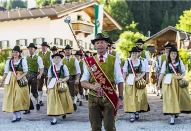 A traditional festival parade with people in traditional costumes. In the foreground, a man with a scepter leads the group.