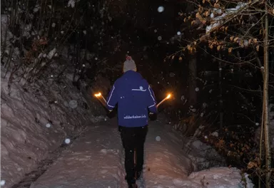 Eine Person geht einen schneebedeckten Weg entlang und hält Fackeln in den Händen. Es schneit und die Umgebung ist dunkel.