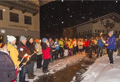 Eine Gruppe von Menschen steht im Schnee und hält Fackeln. Es schneit, und die Szene findet bei Nacht statt.