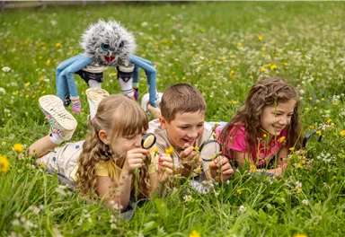 Four children are lying on a meadow and looking at the grass with a magnifying glass. A colorful, creatively designed creature is observing them from behind.