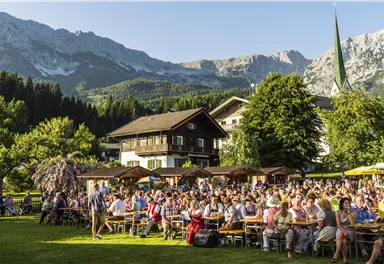 Eine große Menschenmenge sitzt im Freien vor einem schönen Bergpanorama. Im Hintergrund sind traditionelle Gebäude und Bäume zu sehen.