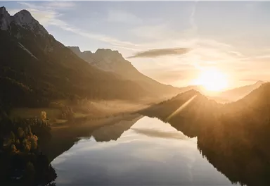 Ein malerischer Sonnenaufgang über einem ruhigen See, umgeben von Bergen. Die sanften Lichtstrahlen spiegeln sich im Wasser wider.