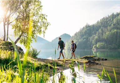 Two hikers are walking along the shore of a clear lake. Surrounded by green trees and hills in the background.