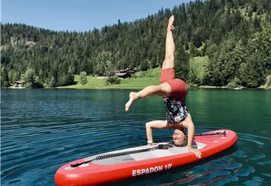 A person is doing a handstand on a stand-up paddleboard in the clear water of a lake. In the background, green trees and rolling landscapes can be seen.