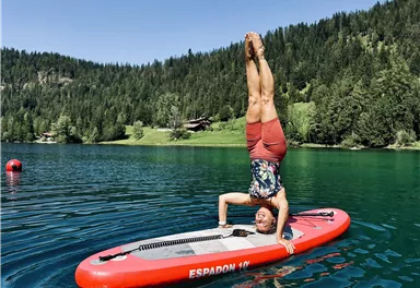 A man is doing a handstand on a stand-up paddleboard on a calm, clear lake. In the background, green trees and a sunny landscape can be seen.