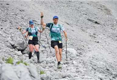Zwei Sportler gehen mit Wanderstöcken über ein felsiges Gelände. Sie tragen leichte Sportkleidung und wirken motiviert.
