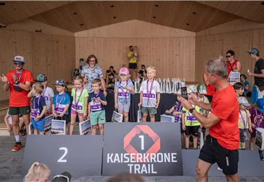 Eine Gruppe von Kindern steht auf einer Bühne beim Kaiserkrone Trail. Sie halten Medaillen und Pokale, während Erwachsene sie anfeuern.
