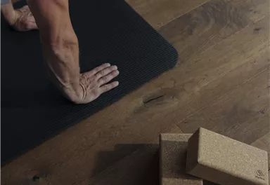 Eine Person macht eine Yoga-Übung auf einer Matte. Neben ihr liegen zwei Korkblöcke auf einem Holzboden.