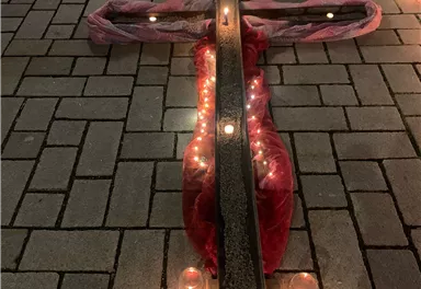 Ein großes Kreuz aus Holz und Stoff liegt auf dem Boden, umgeben von Kerzen. Im Hintergrund sind Lichter und eine Botschaft sichtbar.