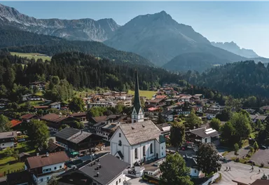 Eine malerische Dorfansicht mit einer weißen Kirche im Vordergrund und den Alpen im Hintergrund. Die Umgebung ist grün und bergig, ideal für Naturliebhaber.