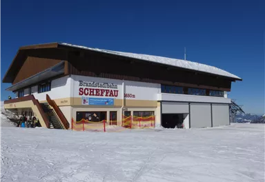 A large building named "Brandstätterbahn Scheffau" in a snowy mountain area. The clear blue sky and the snow create a winter atmosphere.