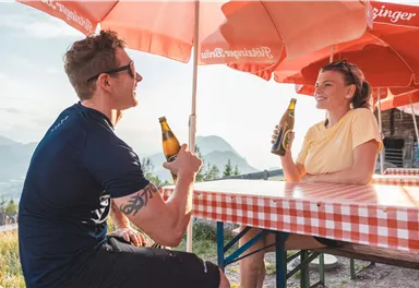 Zwei Personen sitzen unter einem Sonnenschirm und genießen ihre Getränke. Im Hintergrund ist eine wunderschöne Berglandschaft zu sehen.