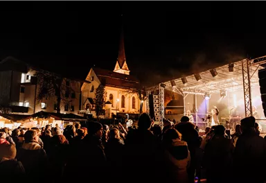 An evening concert in a festively lit city. Many people are standing in front of the stage and enjoying the music.