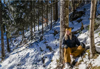 Eine Person sitzt im Schnee, gestützt an einem Baum. Umgeben von verschneiten Bäumen und einer klaren Winterlandschaft.