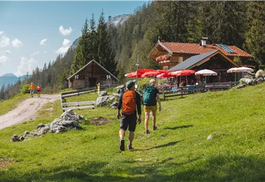 Two hikers are walking on a path to a mountain hut with red sun umbrellas. The surroundings are green and surrounded by trees.