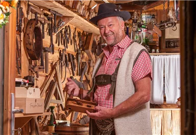 Ein Handwerker steht in seiner Werkstatt und lächelt. Um ihn herum sind viele Werkzeuge und Holzbearbeitungsutensilien.
