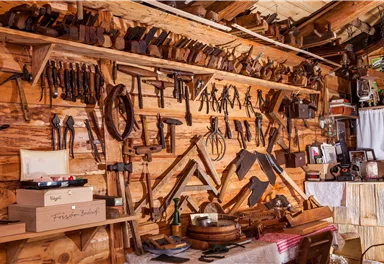 Eine Sammlung von Holzwerkzeugen hängt an einer Wand in einer Werkstatt. Das Holz und die Werkzeuge vermitteln ein handwerkliches Ambiente.