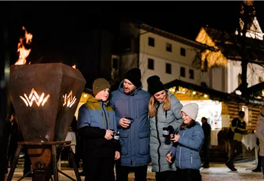 Eine Familie steht in einem festlich beleuchteten Markt und genießt warme Getränke. Im Hintergrund brennen Flammen und es sind festliche Stände zu sehen.