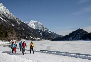 Eine Gruppe von fünf Menschen wandert am Ufer eines zugefrorenen Sees. Im Hintergrund sind schneebedeckte Berge und klare blaue Himmel zu sehen.