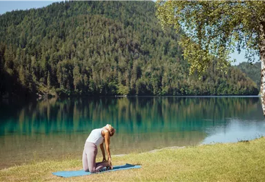 Eine Frau macht Yoga am Ufer eines ruhigen Sees. Im Hintergrund sind grüne Hügel und ein klarer Himmel zu sehen.