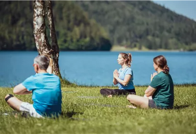 Drei Personen meditieren am Ufer eines Sees. Die Umgebung ist grün und von Bäumen umgeben.