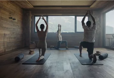 A peaceful yoga class in a wooden room with a view of the mountains. Three people are practicing different yoga poses on mats.