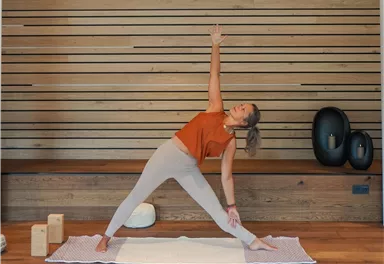 Eine Frau macht Yoga in einem hellen Raum mit Holzverkleidung. Sie befindet sich in der Position des Kriegers und wirkt konzentriert und entspannt.