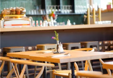 Ein gemütliches Café mit Holztischen und Stühlen. Im Hintergrund sind Getränke und Snacks ausgestellt.
