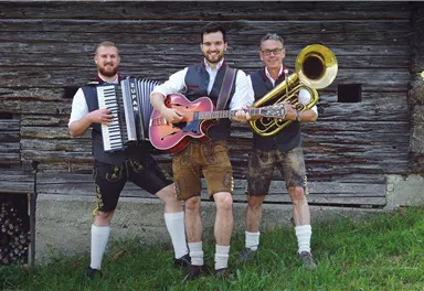 Three musicians in traditional Bavarian attire stand in front of a wooden wall. They are playing an accordion, a guitar, and a tuba.