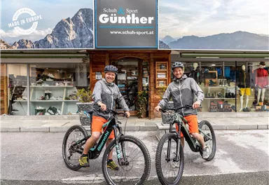 Zwei Radfahrer stehen mit ihren Fahrrädern vor einem Sportgeschäft. Im Hintergrund sind die Berge und das Geschäftsschild sichtbar.