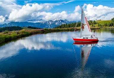 A red sailboat is floating on a calm lake. In the background, there are mountains and a beautiful cloudscape.